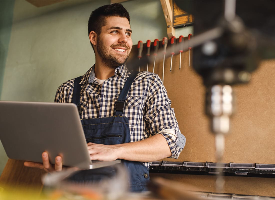 Business Insurance - Portrait of a Smiling Young Carpenter Standing Inside his Workshop While Holding a Laptop